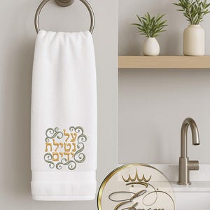 May include: White hand towel with embroidered Hebrew text in gold and green, hanging from a silver towel ring. The towel features a decorative border and is displayed in a bathroom setting.
