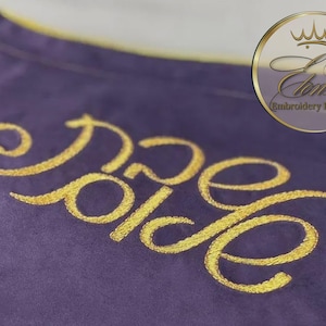 Peut inclure: Gros plan sur un tissu violet avec des lettres brodées en or qui se lisent "Dore Dolce". Le tissu a une bordure dorée. Un logo circulaire avec le texte "L'Consau Embroidery Boutique" est dans le coin supérieur droit.