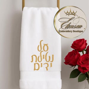 Puede incluir: Toalla de mano blanca con texto hebreo bordado en oro. La toalla está colgada de un toallero dorado. Un ramo de rosas rojas está en un jarrón a la derecha de la toalla. La toalla tiene un borde decorativo.