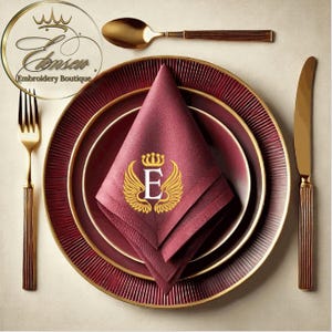 Puede incluir: Una servilleta de lino burdeos con un monograma bordado en oro de la letra "E" con una corona y alas. La servilleta está doblada y colocada sobre un plato de cena burdeos y dorado.