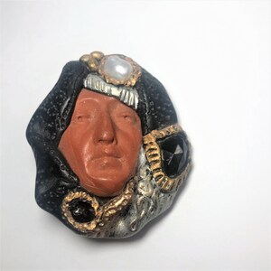 May include: A hand-crafted ceramic brooch featuring a stylized face with a black, gold, and silver design. The face is surrounded by a black fabric-like material and adorned with gold and silver accents. The brooch has a pearl and a black gemstone.