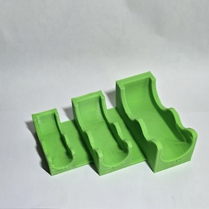 May include: Three bright green, 3D-printed, curved holders of varying sizes. Each holder has a rounded channel and a flat base, connected in a stepped arrangement. The objects are made of plastic and are designed to hold small items.