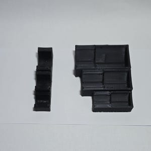 May include: Two black, 3D-printed geometric shapes. One is a tall, narrow, stepped structure. The other is a larger, multi-tiered block with a stepped design. Both are on a white surface.