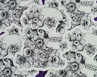 May include: A selection of black and white stickers depicting skulls decorated with flowers. The stickers are set against a purple backdrop. The skulls have detailed features and are surrounded by various types of flowers.