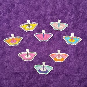 Puede incluir: Siete pegatinas con forma de mariposa dispuestas en grupo. Cada pegatina presenta una combinación de colores única y el texto "WE LOVE DESPITE YOUR HATE" escrito en las alas. Las mariposas muestran tonos amarillos, rosas, azules y naranjas.