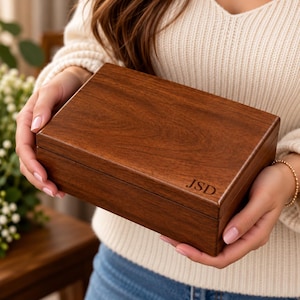 May include: A rectangular wooden box with a smooth, dark brown finish and visible wood grain. The lid features the engraved letters JSD in the bottom right corner. The box has a clean, simple design with straight edges and a hinged construction.