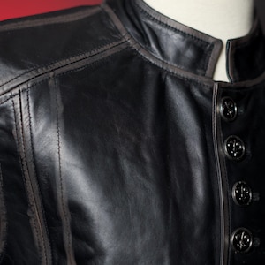 May include: Close-up of a black leather vest with a mandarin collar and decorative buttons. The vest features detailed stitching and a subtle brown trim along the edges, creating a classic and refined look.