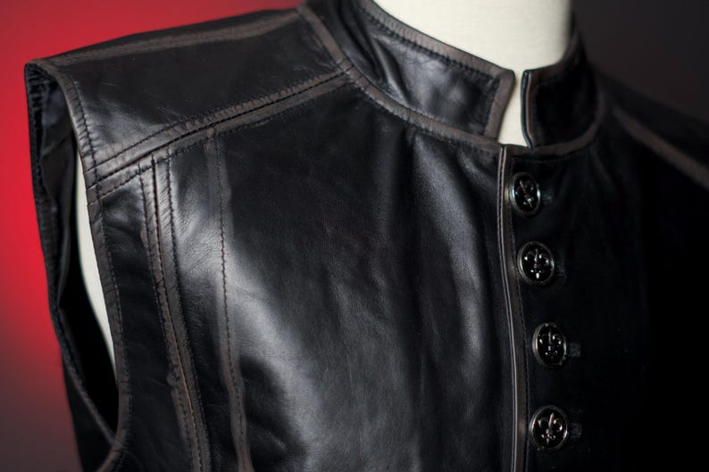 May include: Close-up of a black leather vest with a mandarin collar and decorative buttons. The vest features detailed stitching and a subtle brown trim along the edges, creating a classic and refined look.