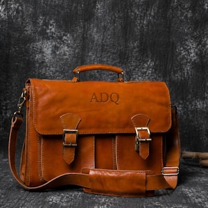 May include: A brown leather satchel with a top handle and shoulder strap. The bag has a flap closure with two buckles and the initials "ADQ" embossed on the front. The bag is made of a rich, textured leather.