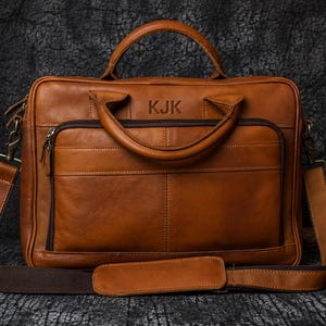 May include: A tan leather briefcase with a top handle and a shoulder strap. The bag has a front pocket with a zip and the initials "KJK" embossed on the top. The bag is on a dark surface.