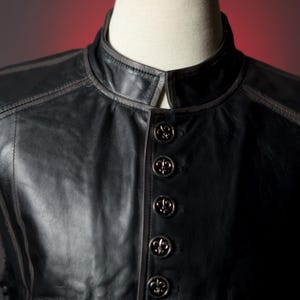 May include: Black leather vest with a mandarin collar and decorative stitching. The vest features five ornate buttons with a fleur-de-lis design. The vest is displayed on a mannequin.