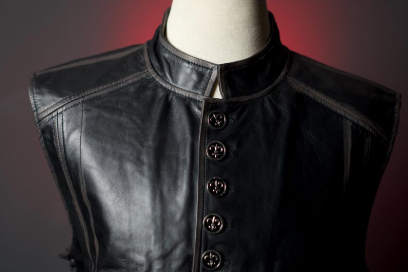 May include: Black leather vest with a mandarin collar and decorative stitching. The vest features five ornate buttons with a fleur-de-lis design. The vest is displayed on a mannequin.