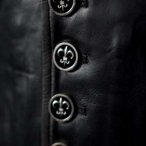 May include: Close-up of a black leather garment with four silver buttons. Each button features a fleur-de-lis design. The buttons are evenly spaced and sewn onto the garment.
