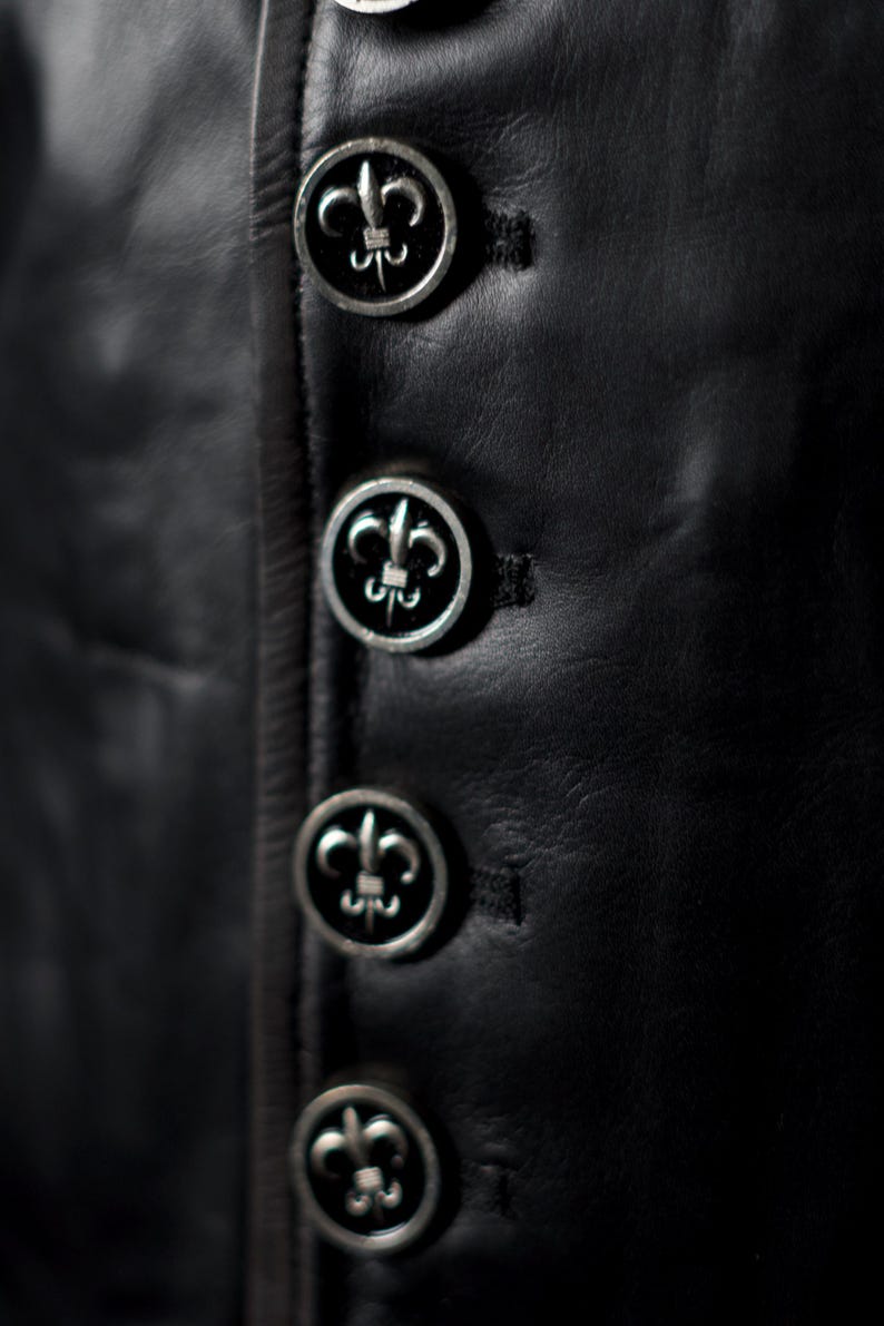 May include: Close-up of a black leather garment with four silver buttons. Each button features a fleur-de-lis design. The buttons are evenly spaced and sewn onto the garment.