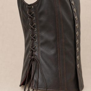 May include: A dark brown leather vest featuring a lace-up side and decorative silver buttons. The vest has a tailored fit and a classic design, suitable for various occasions. The leather appears distressed, adding to its vintage appeal.