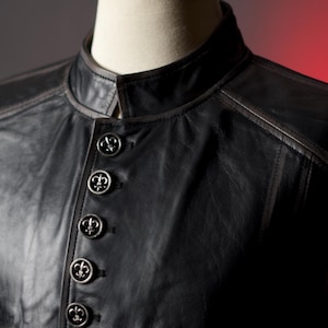 May include: Black leather vest with a mandarin collar and decorative silver buttons. The vest has a tailored fit and detailed stitching. The buttons are embossed with a fleur-de-lis design.