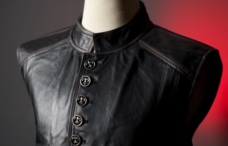 May include: Black leather vest with a mandarin collar and decorative silver buttons. The vest has a tailored fit and detailed stitching. The buttons are embossed with a fleur-de-lis design.
