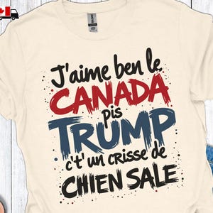 Puede incluir: Camiseta color crema con el texto "J'aime ben le CANADA pis TRUMP c't'un crisse de CHIEN SALE" en negro, rojo y azul. La camiseta tiene cuello redondo y mangas cortas. Una bandera canadiense y un gráfico de camión están en la esquina superior izquierda.