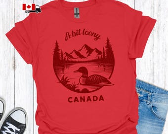 May include: Red t-shirt with a circular graphic featuring a loon on a lake, mountains, and pine trees. Text reads "A bit loony CANADA". The shirt is made of a soft material.