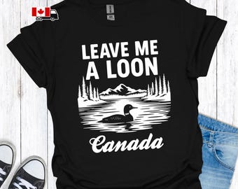 May include: Black t-shirt with a white graphic featuring a loon on a lake, mountains, and trees. The text "LEAVE ME A LOON" and "Canada" are above and below the image. The shirt is displayed on a wooden surface.