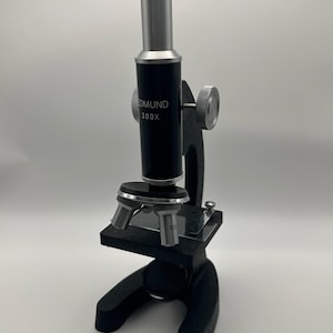 May include: A black and silver Edmund 300X microscope featuring a vertical eyepiece tube, a rotating objective lens turret with a 30X lens, a flat stage with metal clips, and a sturdy base. The main body is marked with the brand name and magnification.