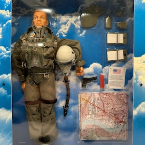 May include: A poseable action figure of a pilot dressed in a grey flight suit and harness, holding a white flight helmet with a communication cord. Accessories include a black handgun, two red flares, a map, and two white rectangular pads.