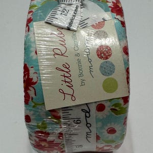 May include: A roll of fabric with a blue and white floral pattern and red accents. The fabric is wrapped in plastic and has a label that says "Little Rubes by Bonnie & Camille" and "moda fabrics.com".