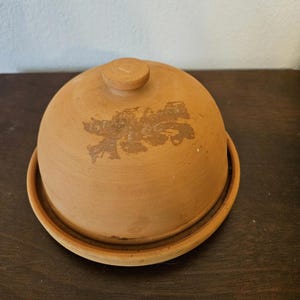 May include: A terracotta cloche with a matching base. The dome-shaped lid has a small knob on top and a faded floral design. The cloche and base are a warm, earthy brown color, perfect for serving or storing food.