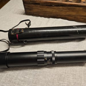 May include: A black telescope with silver accents and a black carrying case with a shoulder strap. The telescope is lying on a light gray surface, with a wooden box in the background.