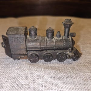 May include: A small, antique-style, dark metal toy train. The train features a detailed design with a smokestack, multiple wheels, and a cabin. The train is sitting on a textured, light-colored surface.