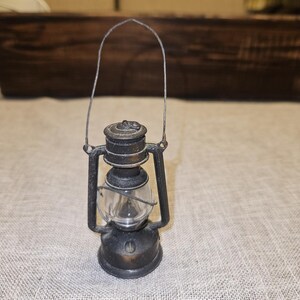 May include: A miniature antique-style lantern with a clear glass globe and a metal frame. The lantern has a handle for carrying and is painted in a dark, aged finish. The lantern is a decorative item.