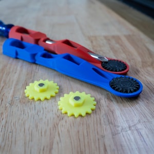 May include: Two blue and red tools with yellow gears on a wooden surface. The tools have a black gear on the end and a handle with a blue or red colour.