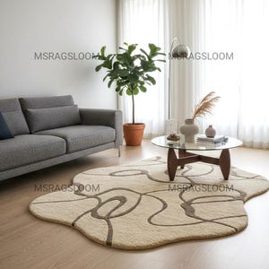 May include: A beige rug with a brown abstract design in a living room. The rug has an unusual shape and is on a wooden floor. A grey sofa, glass coffee table, and a potted plant are also in the room.