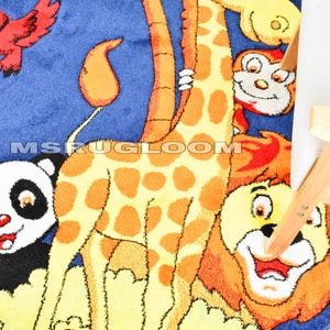 May include: A round children's rug with a blue background and a colorful animal design. The rug features a giraffe, panda, lion, and monkey. The giraffe has orange spots and a long neck. The rug has the text "MS RUG LOOM" on it.