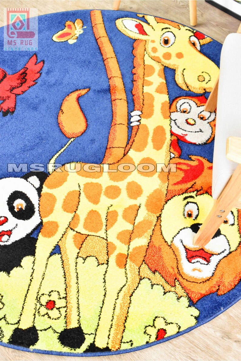 May include: A round children's rug with a blue background and a colorful animal design. The rug features a giraffe, panda, lion, and monkey. The giraffe has orange spots and a long neck. The rug has the text "MS RUG LOOM" on it.