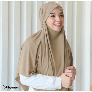 May include: A woman wearing a tan hijab and a white long-sleeved shirt. The hijab is draped over the head and shoulders, with the word "Mocca" in the bottom left corner.