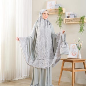 May include: A light gray prayer outfit with intricate white lace detailing. The set includes a matching head covering, a long dress, and a small drawstring bag. The outfit is designed for prayer and is made of a soft, flowing fabric.