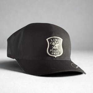 May include: Black baseball cap featuring a Lima Police SWAT emblem. The emblem is a shield shape with the words "LIMA POLICE SWAT" and a graphic. The cap has a curved brim and a structured fit.