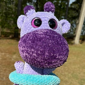 May include: A handmade plush toy in the shape of a purple hippo with large, glittery eyes. The hippo has a dark purple snout, light purple body, and peach-colored legs. A turquoise and blue ring encircles its neck.