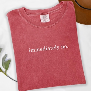 Op de afbeelding: Een rood t-shirt met de witte tekst "immediately no.". Het shirt heeft een ronde hals en is gemaakt van een zacht materiaal. Een bruine hoed is gedeeltelijk zichtbaar op de achtergrond.