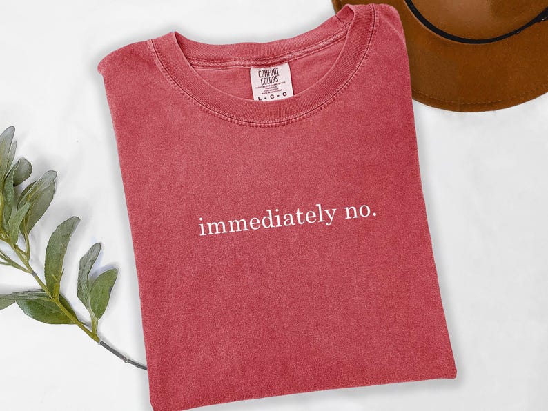 Op de afbeelding: Een rood t-shirt met de witte tekst "immediately no.". Het shirt heeft een ronde hals en is gemaakt van een zacht materiaal. Een bruine hoed is gedeeltelijk zichtbaar op de achtergrond.