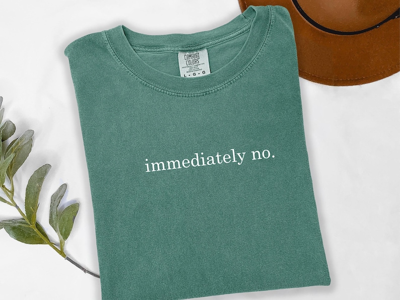 Op de afbeelding: Een teal Comfort Colors t-shirt met de witte tekst "immediately no". Een bruine hoed is zichtbaar in de rechterbovenhoek. Een takje groen is in de linker benedenhoek.
