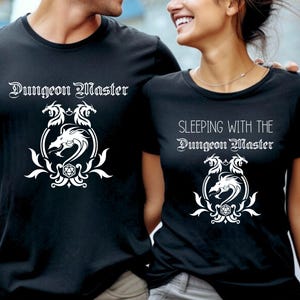 May include: Two black t-shirts with white text and a dragon graphic. The first shirt says "Dungeon Master" and the second shirt says "Sleeping with the Dungeon Master".