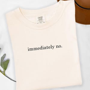 Op de afbeelding: Cr&egrave;mekleurig T-shirt met de zwarte tekst "immediately no.". Het shirt is netjes opgevouwen, met een bruine hoed op de achtergrond. Het shirt heeft een label met "Comfort Colors".