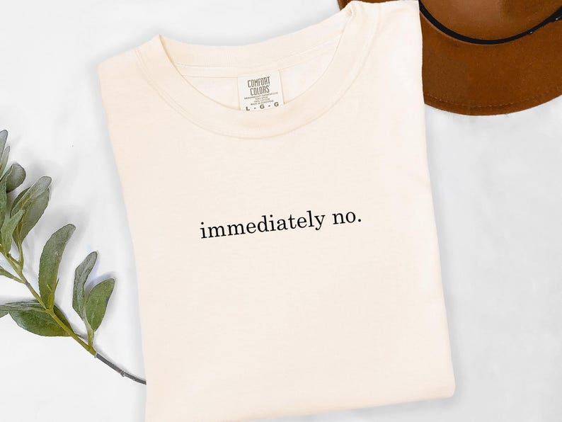Op de afbeelding: Cr&egrave;mekleurig T-shirt met de zwarte tekst "immediately no.". Het shirt is netjes opgevouwen, met een bruine hoed op de achtergrond. Het shirt heeft een label met "Comfort Colors".