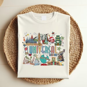 May include: Cream-colored t-shirt featuring a colorful graphic of Universal Studios attractions. The design includes the text "UNIVERSAL STUDIOS" in a rainbow font, along with illustrations of various theme park elements.