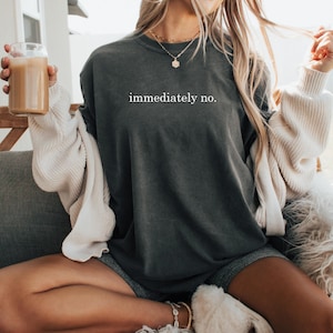 Op de afbeelding: Donkergrijs T-shirt met de witte tekst "immediately no.". Het shirt wordt gedragen met een cr&egrave;mekleurig vest en grijze shorts. Een helder glas met een bruine drank wordt vastgehouden.