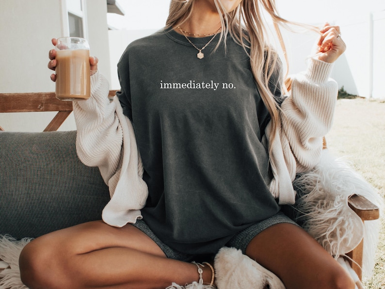 Op de afbeelding: Donkergrijs T-shirt met de witte tekst "immediately no.". Het shirt wordt gedragen met een cr&egrave;mekleurig vest en grijze shorts. Een helder glas met een bruine drank wordt vastgehouden.
