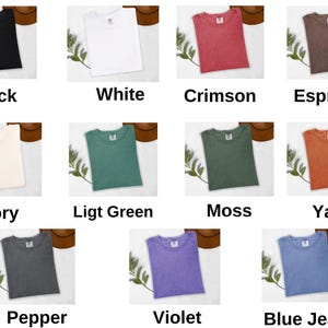 Op de afbeelding: Een verzameling t-shirts in verschillende kleuren, waaronder zwart, wit, karmozijnrood en espresso. Elk shirt wordt gepresenteerd met de kleurnaam eronder. Andere kleuren zijn ivoor, lichtgroen, mos, yam, peper, violet en blue jean.