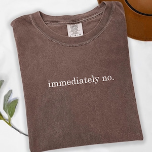 Op de afbeelding: Een bruin Comfort Colors T-shirt met de witte tekst "immediately no.". Het shirt ligt opgevouwen op een witte ondergrond, met een bruine hoed op de achtergrond. Een takje groene bladeren bevindt zich in de linker benedenhoek.